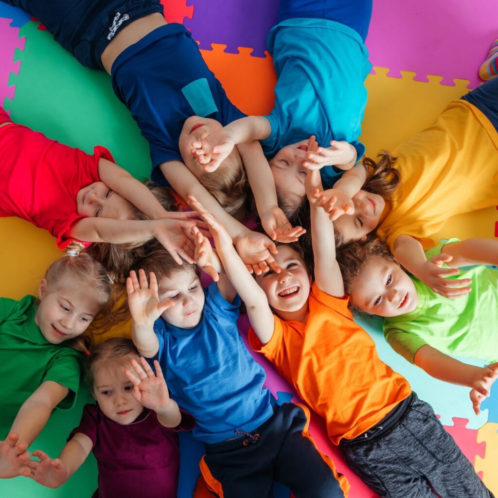 Fröhliche Kinder in bunten T-Shirts liegen lachend auf einer bunten Matte in der Kindertagesstätte SchlaWiener. Optimale Förderung in einem ländlichen Umfeld.